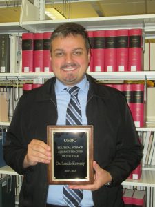 Picture of Laszlo Korossy holding his UMBC plaque award as UMBC's Political Science Adjunct Teacher of the Year dated in 2017-2018. He stands in front of a shelve of red books located in a library.