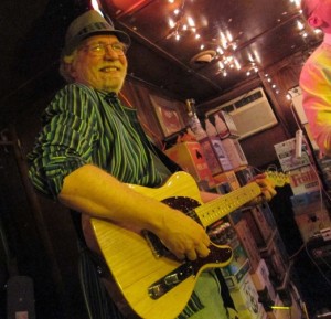 Photo of Timothy Brennan wearing a fedora and green striped shirt, playing stylishly on a Fender Telecaster guitar at a party. There can be white party lights seen attached on the ceiling, with red hue mood lights lighting the room for a fun party atmosphere.