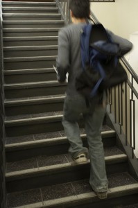 Photo of a student walking upstairs with an unzipped blue backpack slung over his right arm.