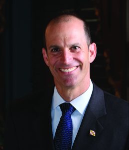 Headshot of Jon Cardin wearing a black suit and blue tie formally patterned with multiple white dots in a grid-like fashion.