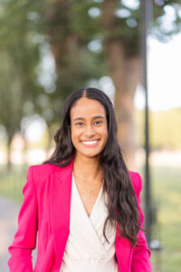 Headshot of woman with long hair and a pink blazer, standing in front of a background of blurry trees